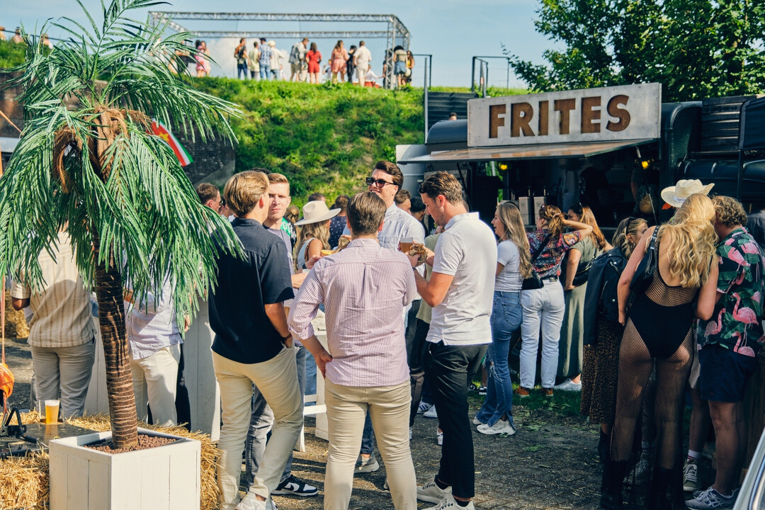 Groep mensen verzameld rond een frietwagen met een groot bord "FRITES" in een buitenomgeving. Sommigen staan te praten en frietjes te eten, terwijl anderen wachten op hun bestelling. De sfeer is zomers en ontspannen, met strobalen en een kunstpalmboom die bijdragen aan een feestelijke setting. Op de achtergrond zijn meer mensen te zien die genieten van het evenement.