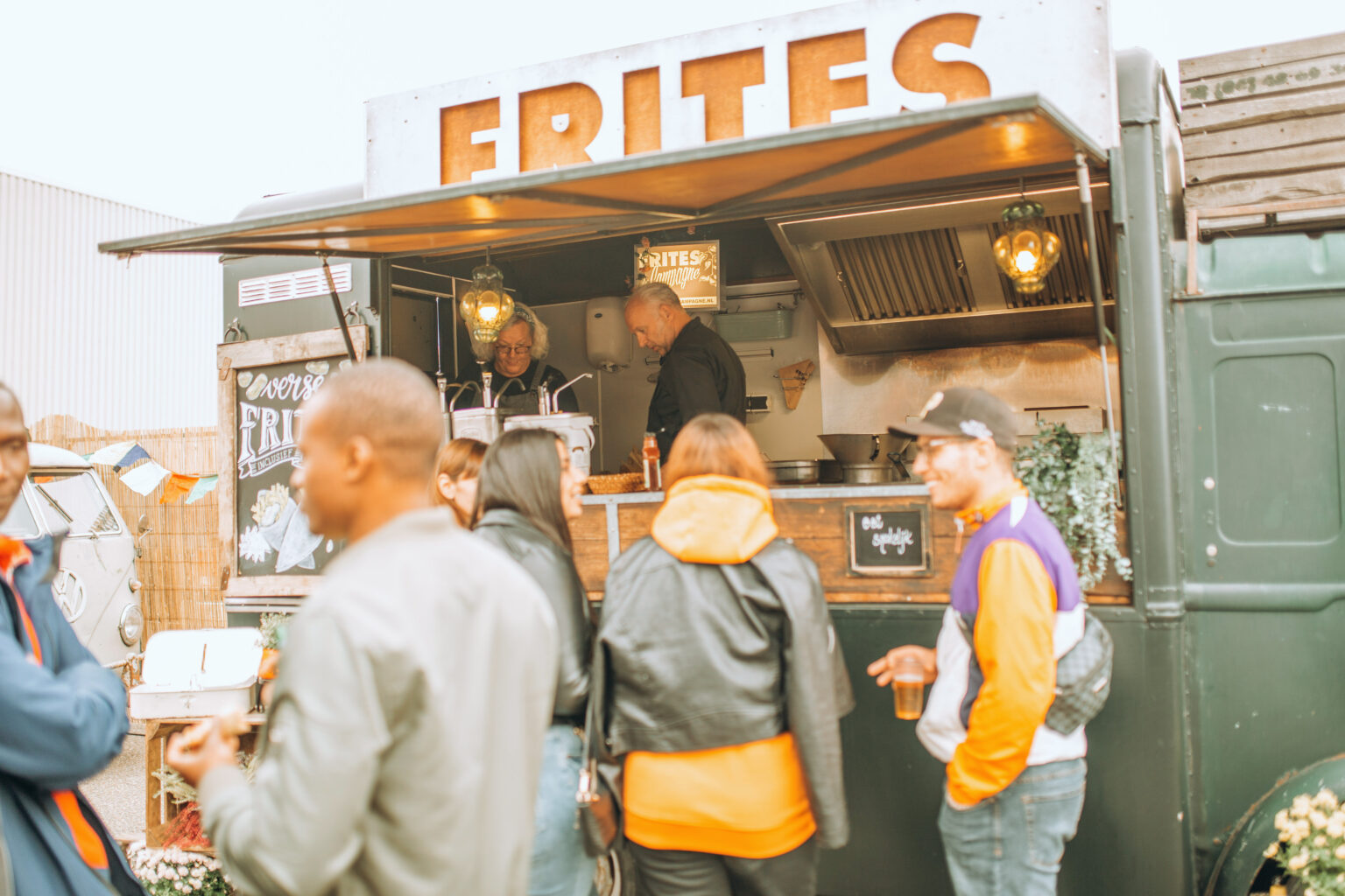 Mensen staan voor een frietwagen met een groot bord "FRITES". De klanten, gekleed in casual jassen, wachten op hun bestellingen terwijl de medewerkers binnen frietjes bereiden. De sfeer is gezellig en ontspannen, met een bordje "Eet smakelijk" op de toonbank en decoratieve elementen rond de wagen.