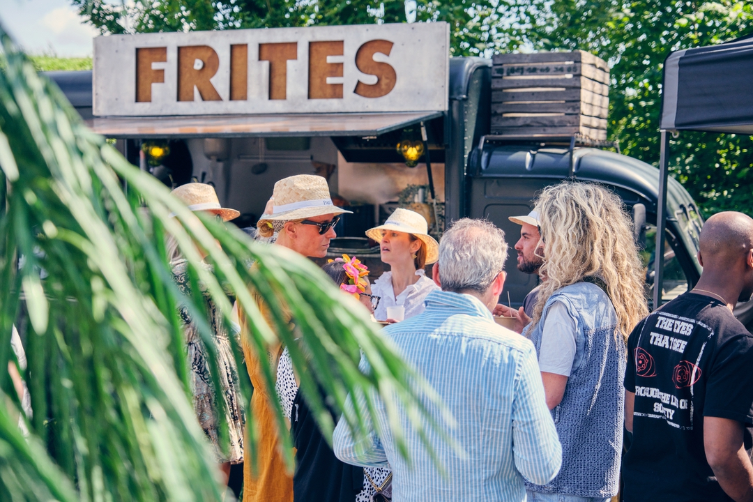 Mensen staan voor een frietkar met een groot bord "FRITES". Ze dragen hoeden en zomerse kleding terwijl ze gezellig praten en frietjes eten. De setting is zonnig met groene bladeren op de voorgrond, wat bijdraagt aan een ontspannen en feestelijke sfeer.