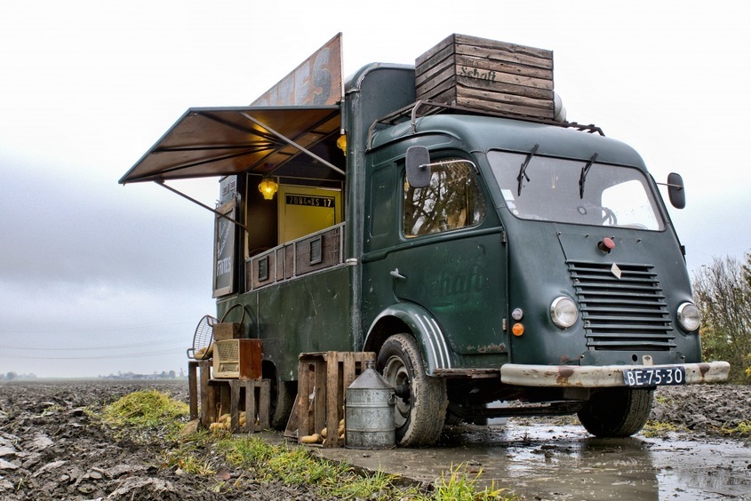 Een vintage groene foodtruck staat op een modderig veld, klaar om friet te serveren. De truck heeft een open luifel en houten kratten op het dak, met een rustieke uitstraling die perfect past bij een landelijke omgeving. Ondanks de grijze lucht en natte omstandigheden, straalt de foodtruck charme en karakter uit.