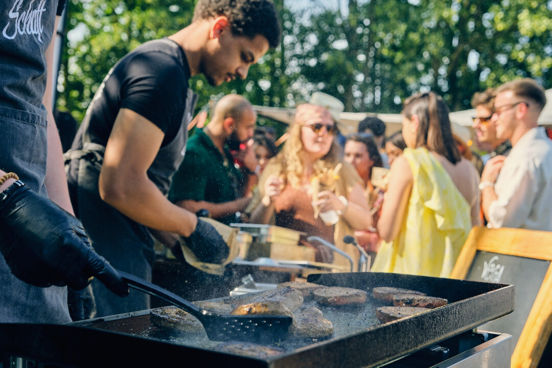 Een kok bakt vlees op een grote grillplaat tijdens een evenement. Op de achtergrond staan mensen in een rij, genietend van hun eten en drinken terwijl ze wachten. De sfeer is levendig en zonnig, met de kok gefocust op het bereiden van de gerechten.