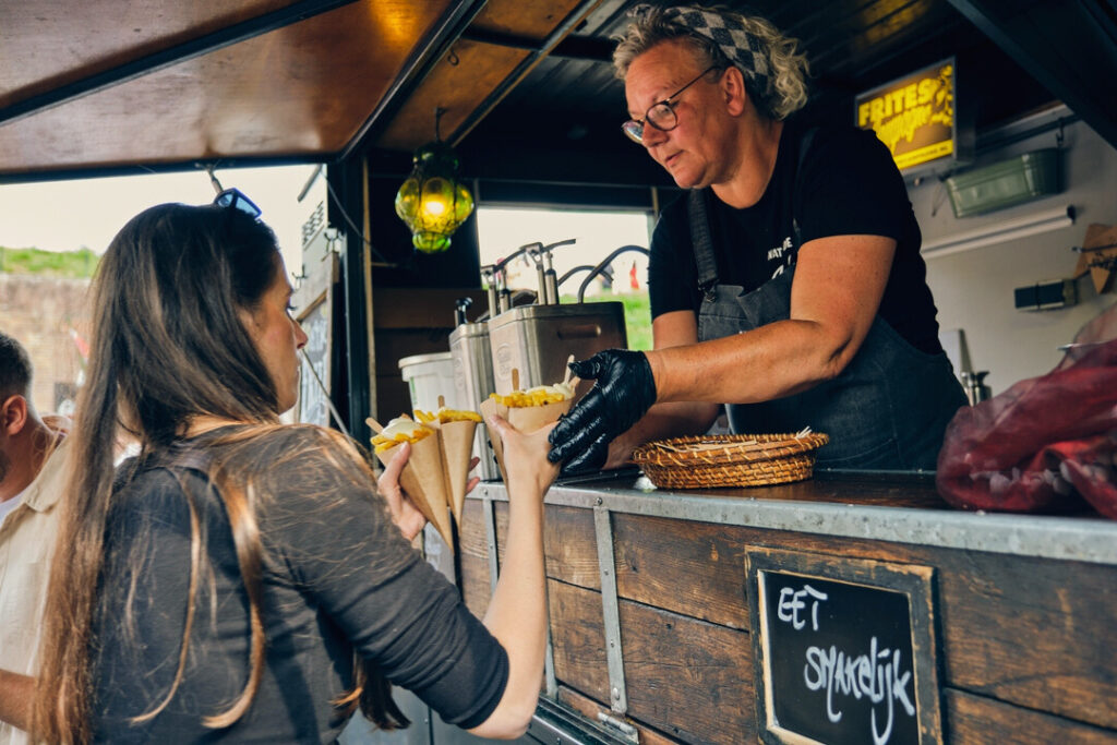 Een klant ontvangt twee porties friet in papieren puntzakken van een medewerker bij een patatwagen. De medewerker, gekleed in een zwart shirt en grijze schort, reikt de frietjes over de toonbank aan. Op een bordje aan de zijkant van de toonbank staat "Eet smakelijk".