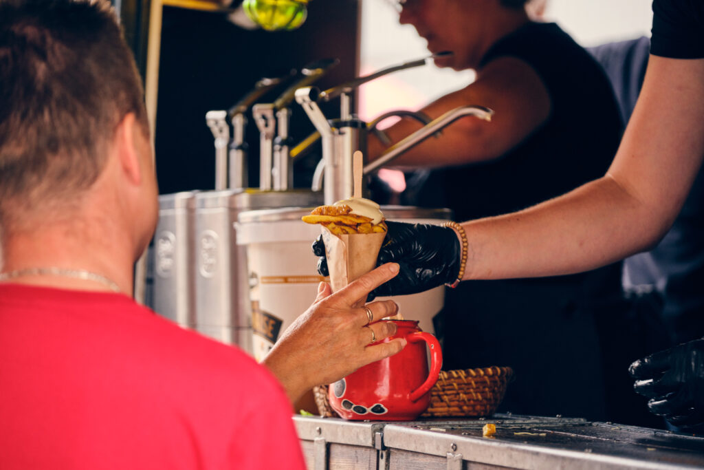 Een klant ontvangt een puntzak friet met mayonaise van een medewerker bij een foodtruck. De medewerker draagt zwarte handschoenen en reikt de frietjes over de toonbank. Op de achtergrond zijn diverse sausdispensers zichtbaar.