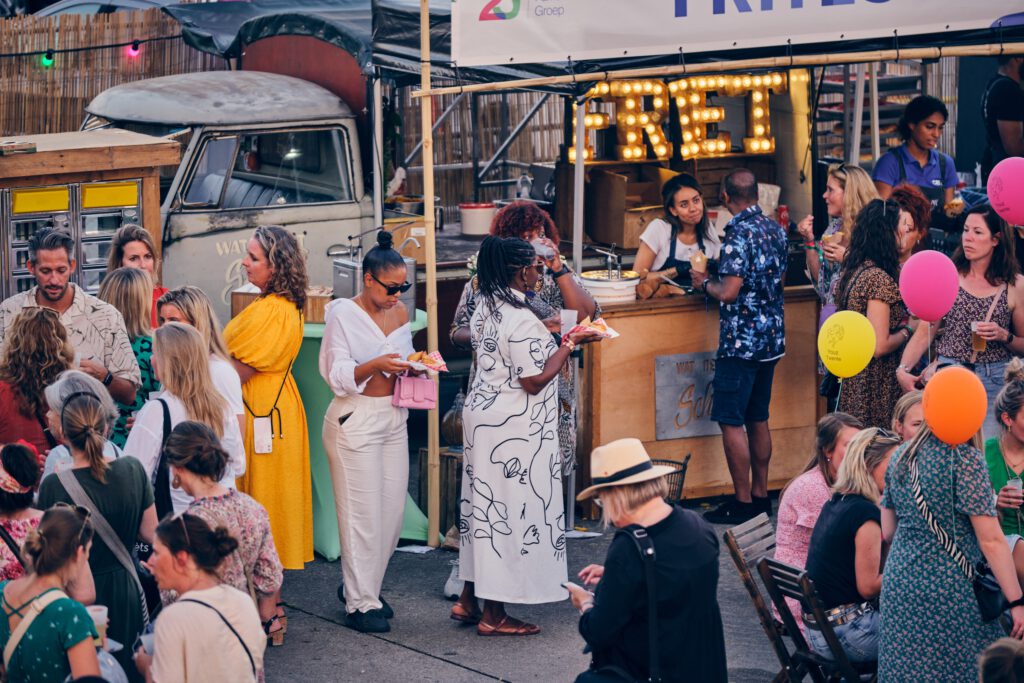 Verschillende mensen staan in de rij voor een foodtruck, zij praten en eten frietjes. De sfeer is levendig met kleurrijke kleding, ballonnen en een vintage foodtruck op de achtergrond.