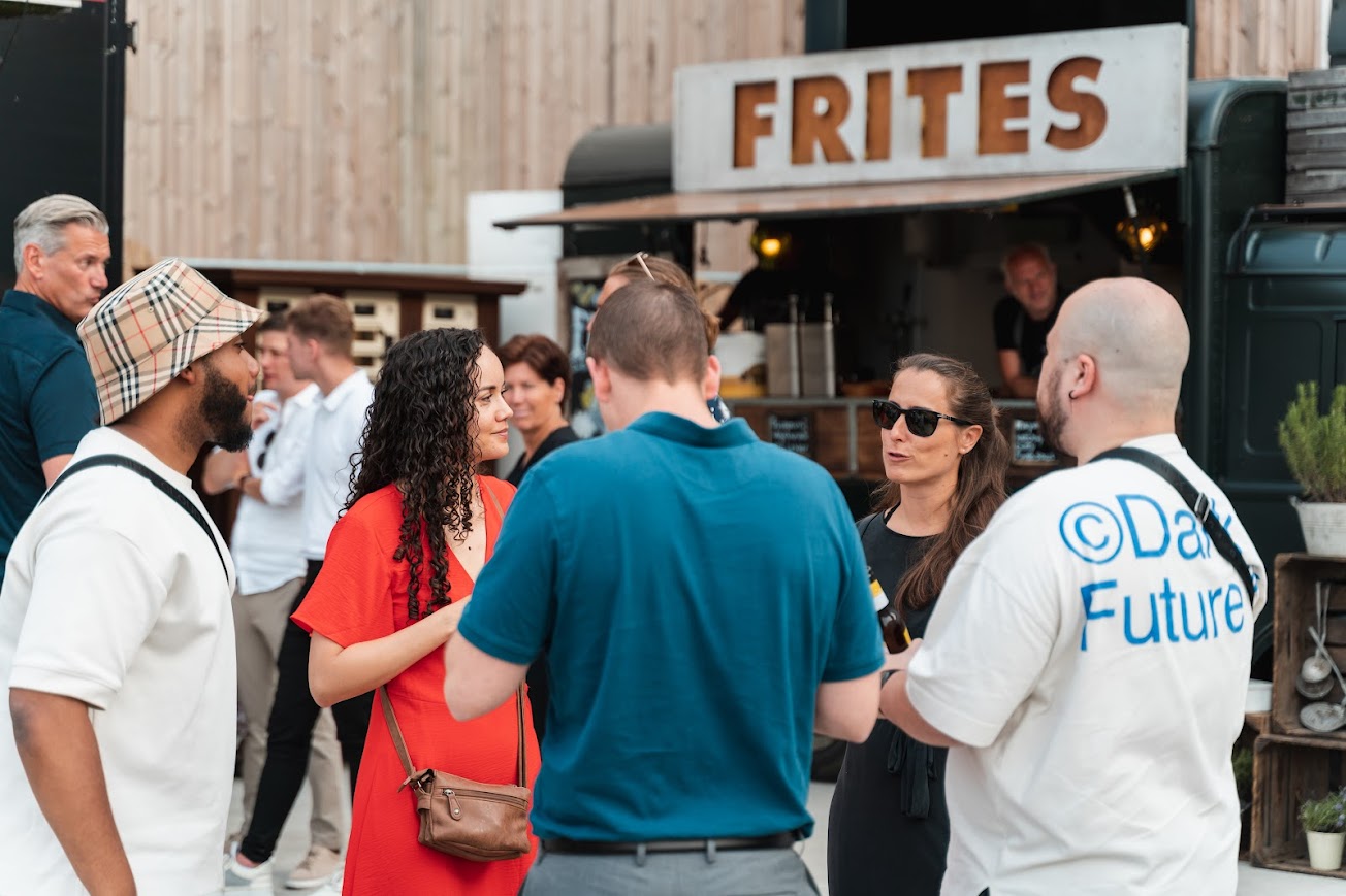 Groep mensen staat voor een foodtruck met een bord "FRITES" erop. Ze praten en lachen terwijl ze genieten van hun eten. De sfeer is ontspannen en sociaal, met verschillende personen in kleurrijke kleding en zomerse accessoires.